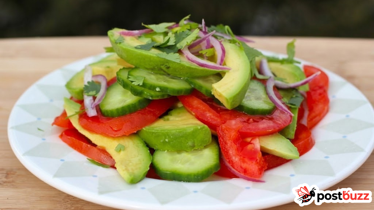 A close-up shot of a fresh Colombian salad served on a white plate with a geometric pattern. The salad is neatly arranged with vibrant layers of sliced ripe tomatoes, crisp cucumber rounds, creamy avocado wedges, and thin slivers of red onion, all topped with freshly chopped cilantro.